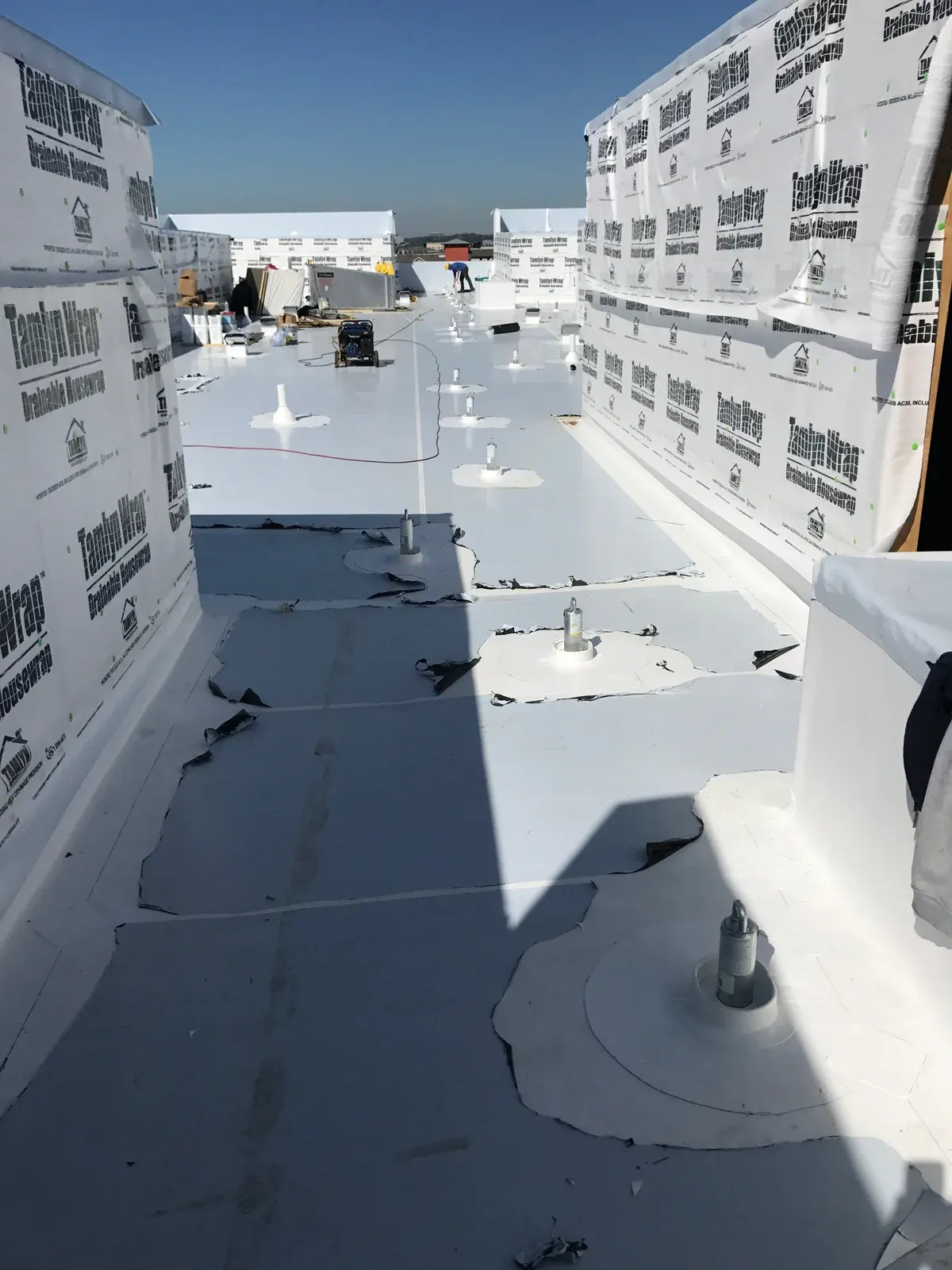 White TPO commercial roof installation in progress showing fastener plates, insulation boards and TamlynWrap wall underlayment with a crew member in the distance